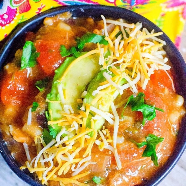 A bowl of Instant Pot turkey soup topped with sliced avocado, shredded cheese, and chopped cilantro. The bowl rests on a wooden surface next to a colorful, patterned fabric. The soup features chunks of vegetables in a rich tomato-based broth.