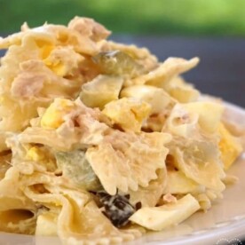 A plate of tuna macaroni salad with bowtie pasta, diced pickles, and boiled egg sits against a blurred green background.