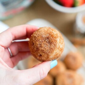 A hand holds a round, cinnamon-sugar-coated treat, reminiscent of pressure cooker churro bites. In the blurred background, more of these tasty delights sit alongside a bowl brimming with strawberries.