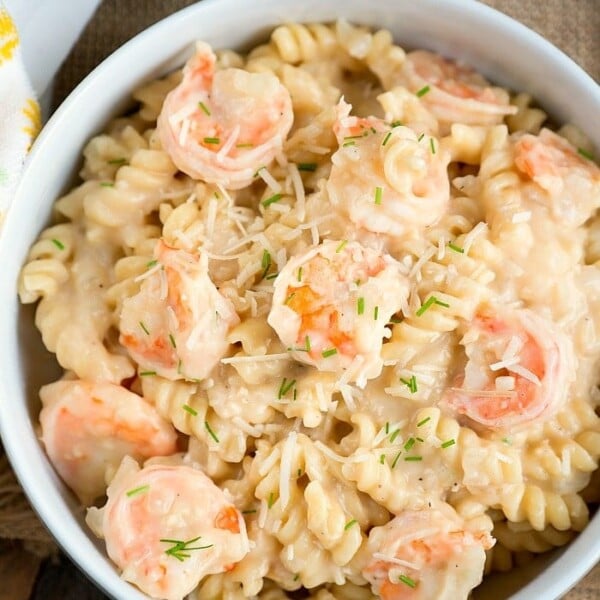 A bowl of creamy instant pot shrimp Alfredo with spiral-shaped noodles, garnished with grated cheese and small bits of herbs. The dish is placed on a wooden surface with a napkin partially visible to the side.