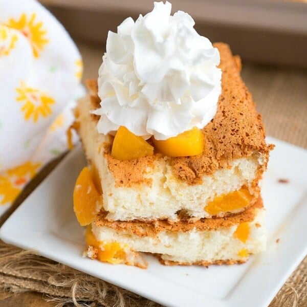 A slice of delightful peach angel food cake topped with whipped cream sits gracefully on a white square plate. The light, fluffy cake reveals juicy peach chunks. A patterned cloth accentuates the scene alongside a baking dish, creating an inviting and warm presentation.