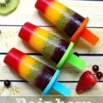 Three vibrant rainbow popsicles with red, orange, yellow, and green layers rest on a wooden background. They have colorful holders in blue, orange, and green. Slices of kiwi, strawberries, and berries surround the popsicles in a delightful array.