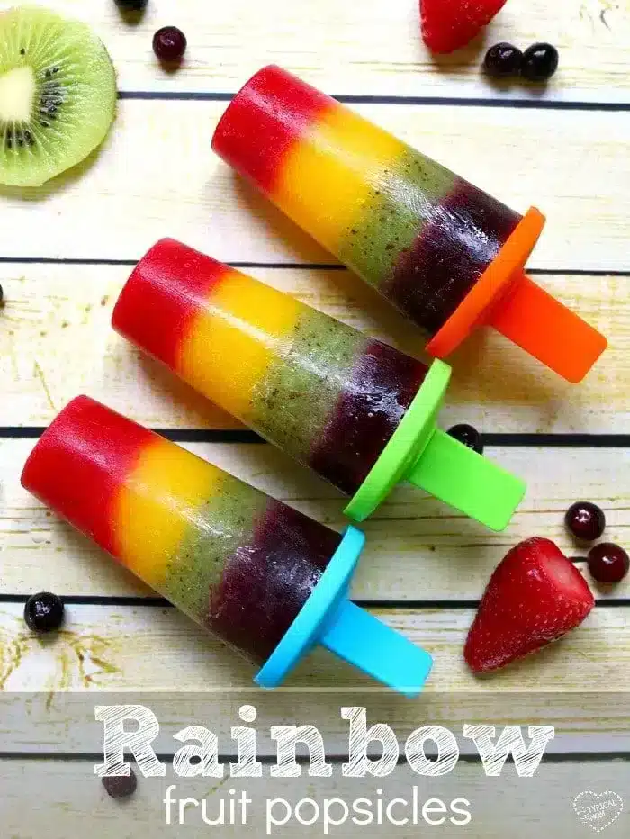 Three vibrant rainbow popsicles with red, orange, yellow, and green layers rest on a wooden background. They have colorful holders in blue, orange, and green. Slices of kiwi, strawberries, and berries surround the popsicles in a delightful array.