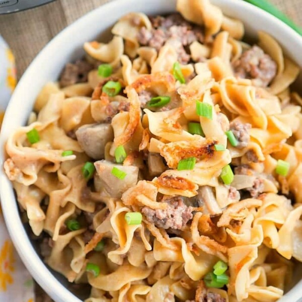 A bowl filled with creamy stroganoff pasta, ground meat, and topped with chopped green onions sits next to a yellow floral-patterned napkin. Nearby, a pressure cooker is partially visible in the background.