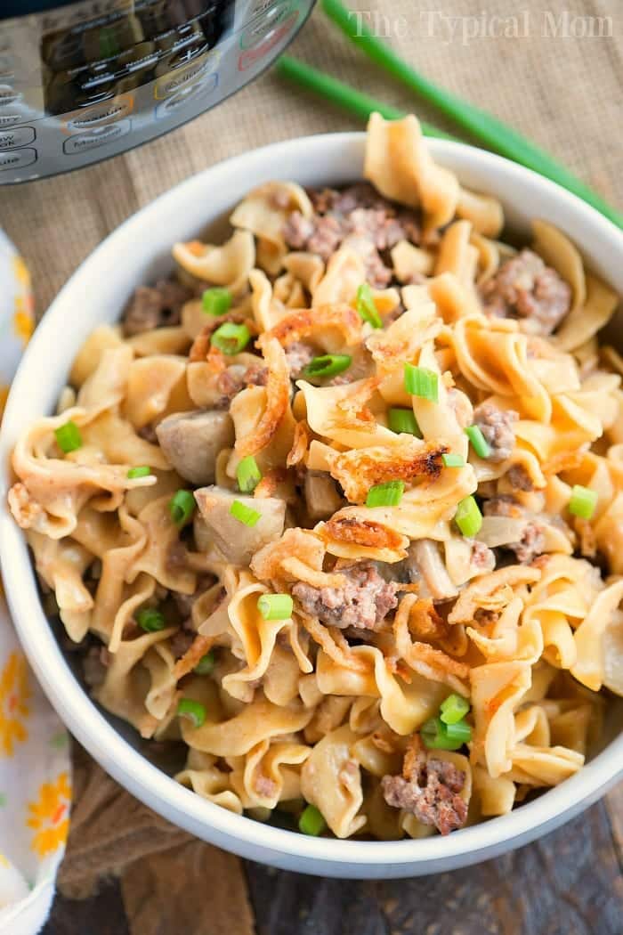 A bowl filled with creamy stroganoff pasta, ground meat, and topped with chopped green onions sits next to a yellow floral-patterned napkin. Nearby, a pressure cooker is partially visible in the background.