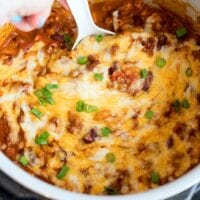 A hand stirs a pressure cooker cheesy chili mac dish, topped with melted cheese and vibrant green onions.