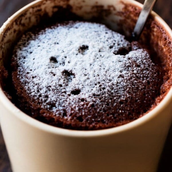 Chocolate mug cake recipe with powdered sugar and a spoon, served in a beige mug on a wooden surface.