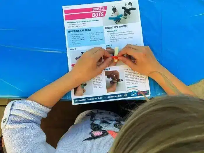 A person is assembling a small project using a guide labeled "Bristle Bots." The guide lists materials and steps. The person, wearing a dog-patterned sweater, is working on a blue tablecloth reminiscent of crafts at vacation Bible school in Temecula.