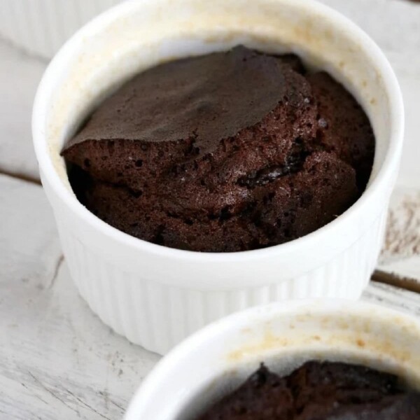 A chocolate soufflé in a white ramekin rests elegantly on the wooden table, its slightly crispy and cracked top reminiscent of rustic charm, surrounded by dishes echoing the warmth of a crockpot cake.