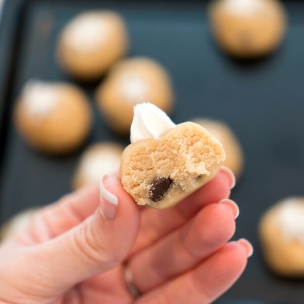 Close-up of a hand holding a half-eaten ball of edible raw cookie dough with a chocolate chip peeking out. Other dough balls and a baking tray are blurred in the background, tempting you with their gooey promise.