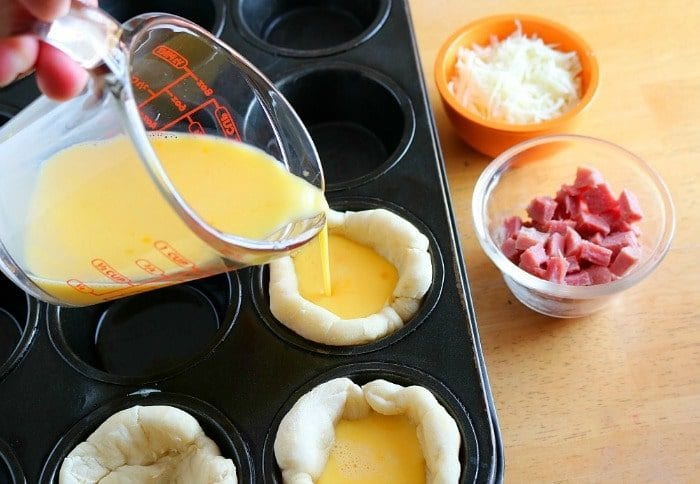 Pouring egg mixture into dough cups in a muffin tin, creating delightful ham and cheese biscuits with bowls of cheese and ham nearby.