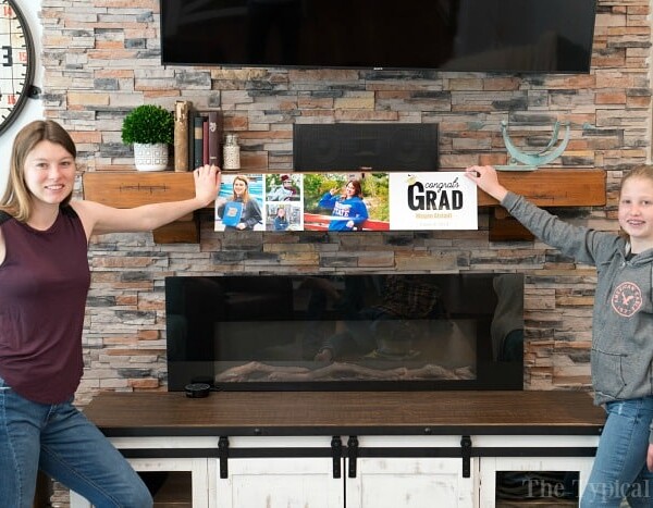 Two girls proudly showcase creative graduation banner ideas indoors, standing by a stone fireplace.