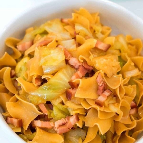 A bowl of egg noodles, infused with pressure cooker cabbage and pieces of cooked bacon. The noodles are golden, and the cabbage is lightly sautéed. This comforting dish is served in a white bowl resting on a light-colored surface.