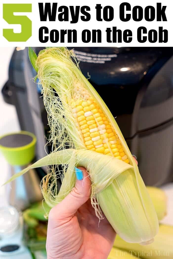 A hand holding an ear of corn partially husked with a blurred kitchen background. Text at the top reads: "5 Ways to Cook Corn on the Cob, including Air Fryer Corn.