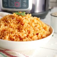 A bowl of vibrant, orange-hued Mexican rice garnished with a sprig of cilantro rests on a wooden table. Nearby, a colorful towel adds charm, while a pressure cooker sits in the background, hinting at the flavorful journey this dish has taken.