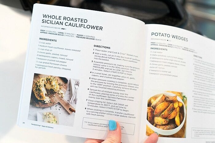 A hand holds open a cookbook, displaying recipes for roasted cauliflower&mdash;including whole pressure cooker cauliflower&mdash;and potato wedges alongside vibrant recipe photos.