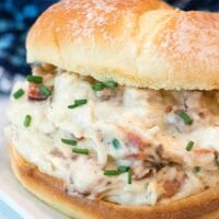 A sandwich filled with creamy Ninja Foodi crack chicken and bacon, garnished with chopped chives. It sits on a square, white plate, with a blurred blue patterned background.