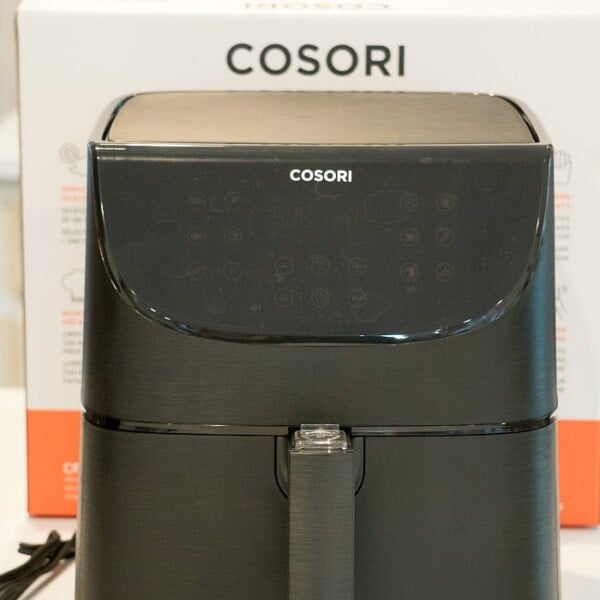 A sleek black electric air fryer by Cosori sits on the kitchen counter, accompanied by its packaging box in the background. The air fryer features a digital control panel on the front, while a power cord is visible to the left.