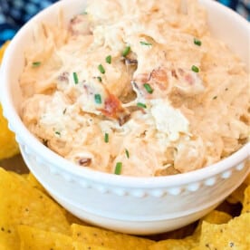 A white bowl brimming with creamy pressure cooker crack chicken dip, garnished with chopped chives. The bowl is surrounded by yellow tortilla chips on a table, inviting you to dive into this delicious creation.