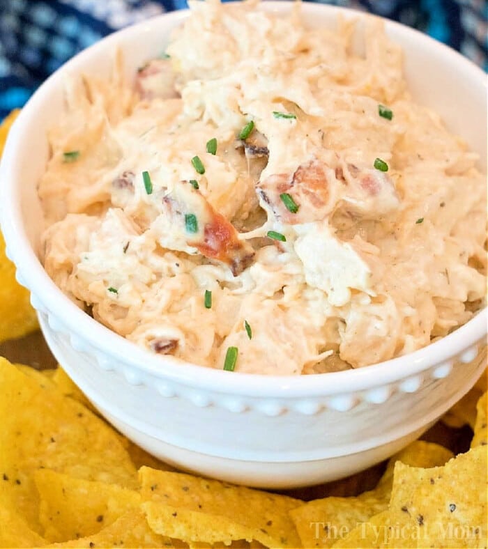 A white bowl brimming with creamy pressure cooker crack chicken dip, garnished with chopped chives. The bowl is surrounded by yellow tortilla chips on a table, inviting you to dive into this delicious creation.