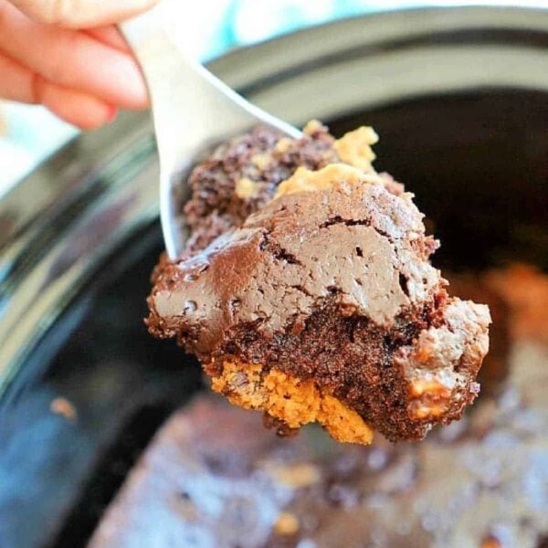 Close-up of a hand holding a spoonful of rich, gooey chocolate cake from a crockpot. The cake appears moist, with a fudgy texture reminiscent of brownie cookies and crispy edges.