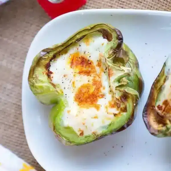 A halved green bell pepper filled with a baked egg sits on a white plate, having been perfectly cooked in an air fryer. The egg is golden brown on top. The background includes a burlap fabric and a colorful napkin with floral patterns, making these breakfast stuffed peppers truly inviting.