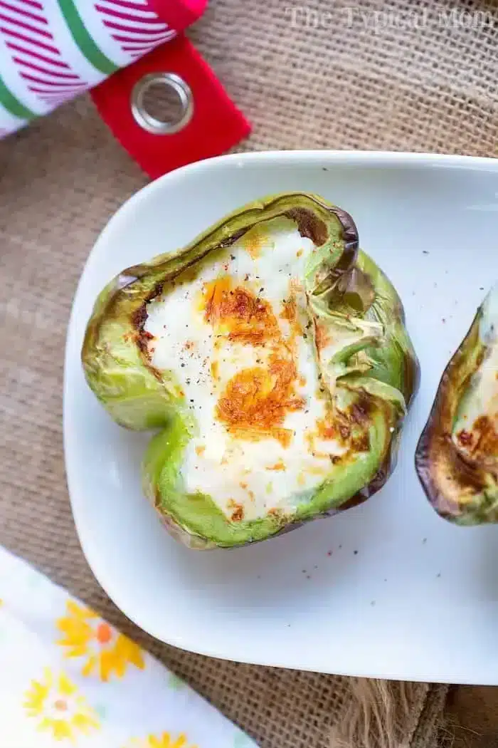 A halved green bell pepper filled with a baked egg sits on a white plate, having been perfectly cooked in an air fryer. The egg is golden brown on top. The background includes a burlap fabric and a colorful napkin with floral patterns, making these breakfast stuffed peppers truly inviting.