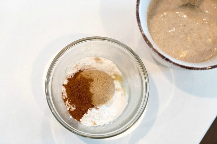 Glass bowl with flour, sugar, and cinnamon sits beside a mug containing a mixed, beige batter with a spoon&mdash;perfect for an easy mug cake recipe.