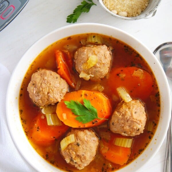 A white bowl contains instant pot meatball soup with sliced carrots and celery, garnished with a parsley leaf. A spoon is on the side, and a small bowl with grated cheese is in the background on a white surface.