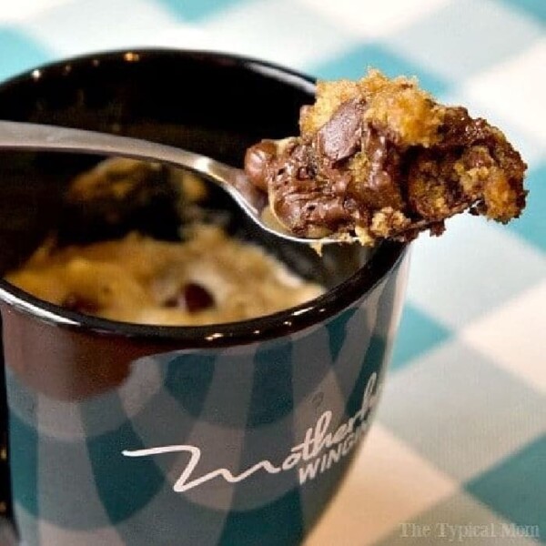 A spoon filled with chocolate chip cookie dough hovers over a black mug, invitingly set on a checkered tablecloth, promising the perfect chocolate chip cookie in a mug.