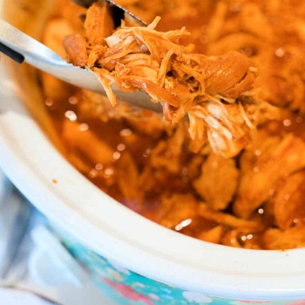 Close-up of pulled chicken being held by tongs above a crockpot. This BBQ chicken recipe showcases tender, shredded meat in a savory sauce, with the slow cooker lined with a floral pattern on the outside.