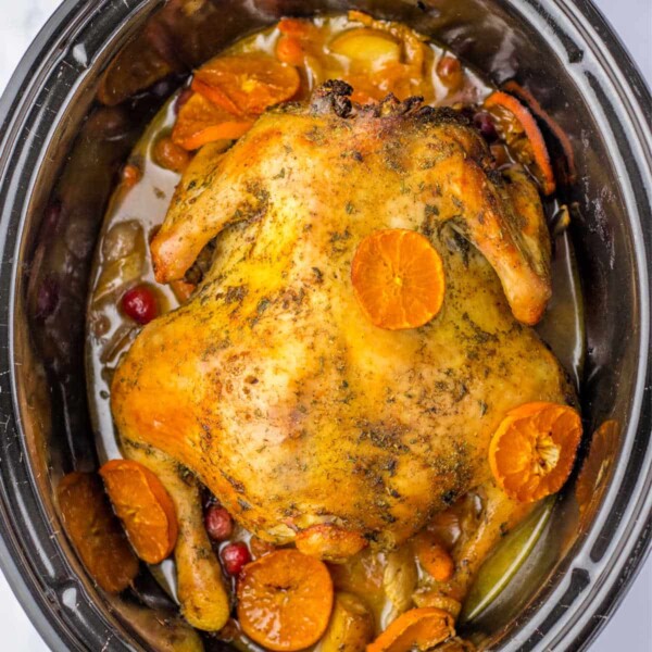 A whole chicken crockpot with potatoes, nestled among sliced carrots, baby potatoes, and juicy oranges. This vibrant dish slow-cooks to perfection in a flavorful broth speckled with tangy cranberries.