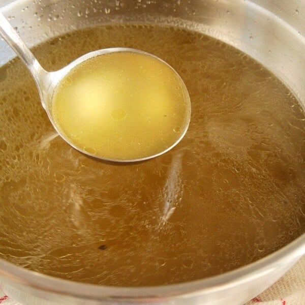 A stainless steel pot, resembling an Instant Pot, is filled with clear chicken broth. A metal ladle hovers above, partially filled with the savory liquid. The pot rests serenely on a cloth surface.