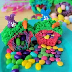 Colorful Surprise Inside Cupcakes with green frosting, topped with plastic bunnies and carrots, cut open to reveal candy treasures inside, displayed on a turquoise plate surrounded by scattered candies.