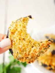 A hand holds a crescent-shaped, golden-brown slice of eggplant, freshly crisped as an air fryer eggplant chip, with a bite taken out.