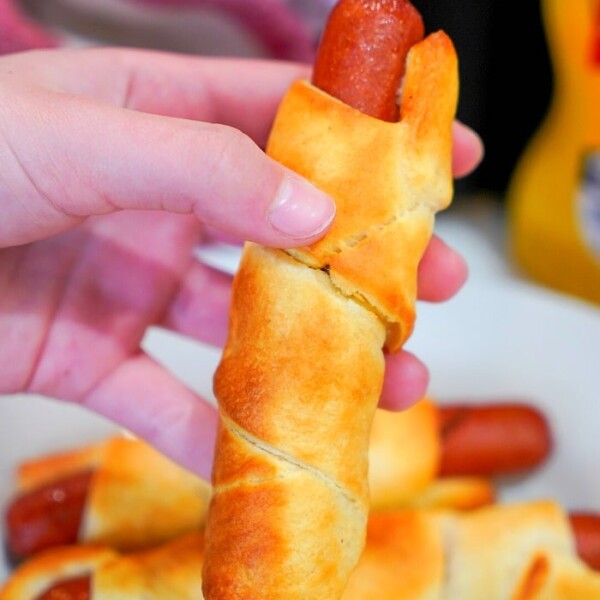 A hand holds a baked crescent roll wrapped around a hot dog, reminiscent of classic air fryer pigs in a blanket. In the background, more wrapped hot dogs rest on a white plate. A bottle of mustard is partially visible to the right.