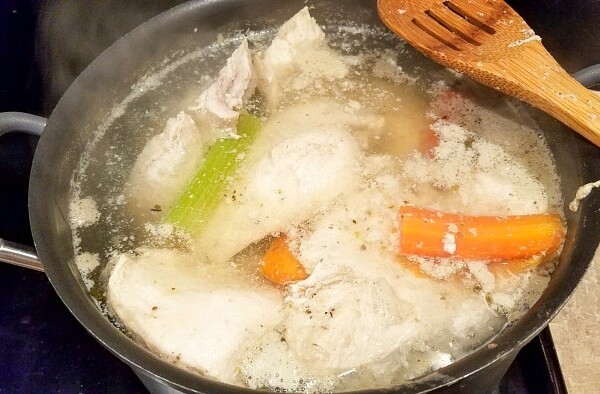 A pot of boiling water with chicken pieces, carrots, and celery simmers on the stove—perfect for learning how to make shredded chicken. A wooden spatula rests on the edge, as steam rises from the bubbling broth.