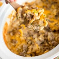 A hand holds a wooden spoon above a crockpot filled with a cheesy hamburger casserole. This savory delight combines macaroni, ground beef, and melted cheese, all garnished with fresh herbs for that perfect finish.