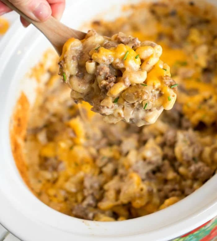 A hand holds a wooden spoon above a crockpot filled with a cheesy hamburger casserole. This savory delight combines macaroni, ground beef, and melted cheese, all garnished with fresh herbs for that perfect finish.