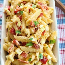 Creamy pasta salad with mayo, penne, diced tomatoes, bacon, and herbs in a rectangular dish on a checkered cloth.