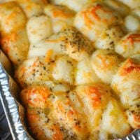 A close-up of a baked dish in a foil tray, showcasing golden-brown smoked bread covered with melted cheese and sprinkled with herbs. The bread appears fluffy and slightly crispy on top, with unevenly distributed cheese and herbs.