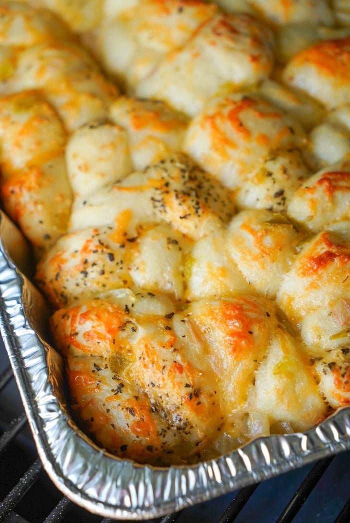 A close-up of a baked dish in a foil tray, showcasing golden-brown smoked bread covered with melted cheese and sprinkled with herbs. The bread appears fluffy and slightly crispy on top, with unevenly distributed cheese and herbs.