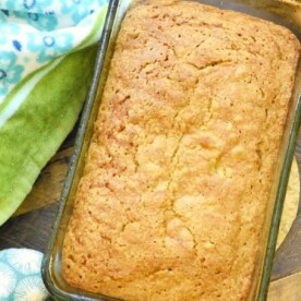 A loaf of banana zucchini bread sits in a glass baking dish on a wooden surface. A green and blue patterned cloth is partially visible in the background. The bread boasts a golden-brown crust with visible cracks on top, hinting at its moist interior.