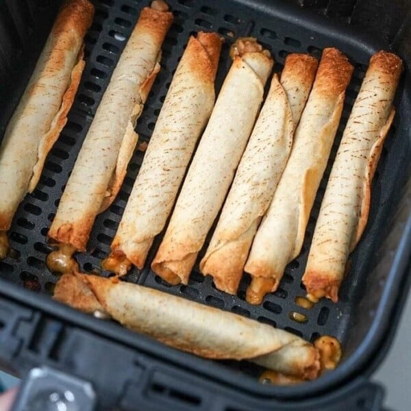 An air fryer basket contains several frozen taquitos arranged in neat rows, now transformed into crispy delights. Each taquito is golden brown with some browned edges, and a bit of cheese has melted out from the ends.