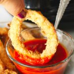 A hand dipping a golden-brown, air fryer calamari ring into a bowl of red sauce.