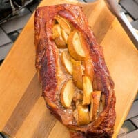 Roasted pork loin stuffed with apple slices on a wooden cutting board, accompanied by tongs, showcases an exquisite smoked pork recipe.