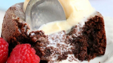 A spoon is cutting into an air fryer chocolate lava cake topped with melting vanilla ice cream on a white plate. Two fresh raspberries sit beside the cake.