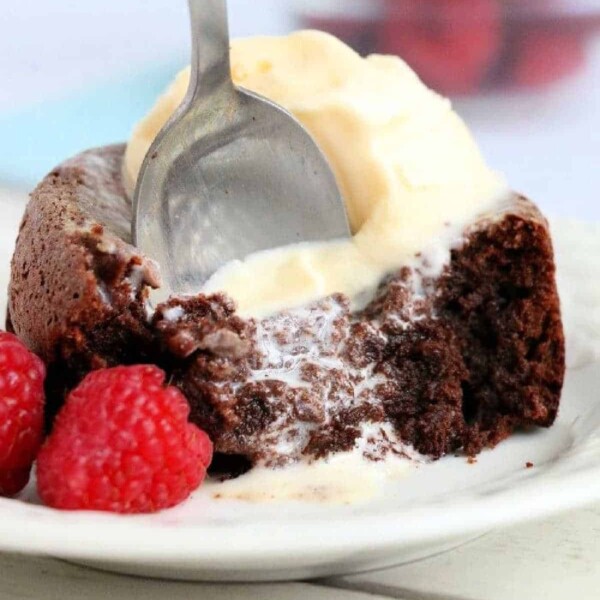 A spoon is cutting into an air fryer chocolate lava cake topped with melting vanilla ice cream on a white plate. Two fresh raspberries sit beside the cake.