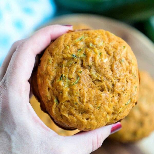 A hand holds a freshly baked pumpkin zucchini muffin over a wooden surface, with another muffin blurred in the background.