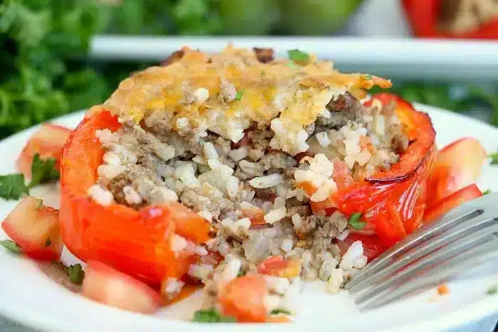 A stuffed red bell pepper, blanched to perfection, sits on a white plate filled with a savory mix of ground meat and rice, topped with melted cheese. Diced tomatoes and chopped herbs are artfully scattered around the pepper, while a fork rests beside it.